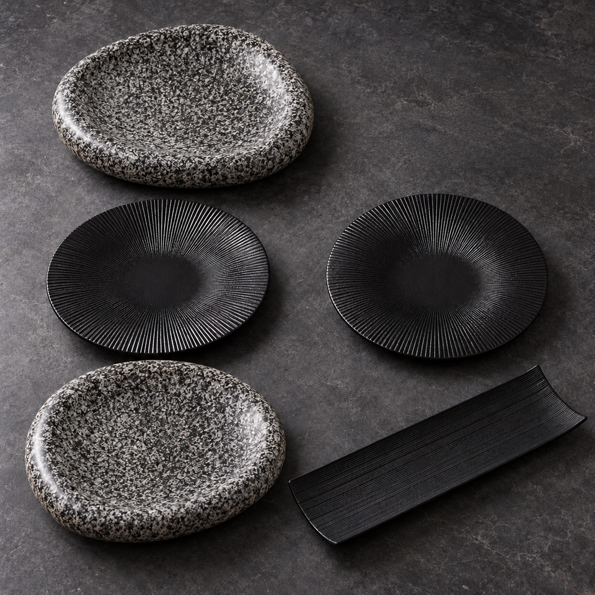 Ensemble de deux assiettes mouchetées, deux petites assiettes noires striées et un plat rectangulaire noir sur fond sombre.