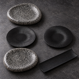 Ensemble de deux assiettes mouchetées, deux petites assiettes noires striées et un plat rectangulaire noir sur fond sombre.
