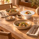 Table en bois avec deux assiettes creuses beiges, une coupe blanche sur pied, une petite assiette et une saucière beige.